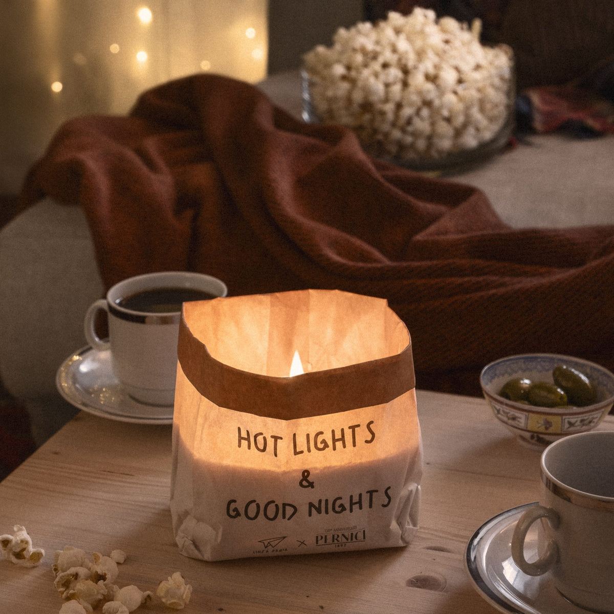 Candela profumata “Hot Lights & Good Nights” accesa in un sacchetto di carta che diffonde una luce calda. La candela è appoggiata su un tavolino in legno con tazze di caffè, popcorn e olive, sullo sfondo una coperta rossa e lucine soffuse che creano un’atmosfera accogliente e rilassata.
