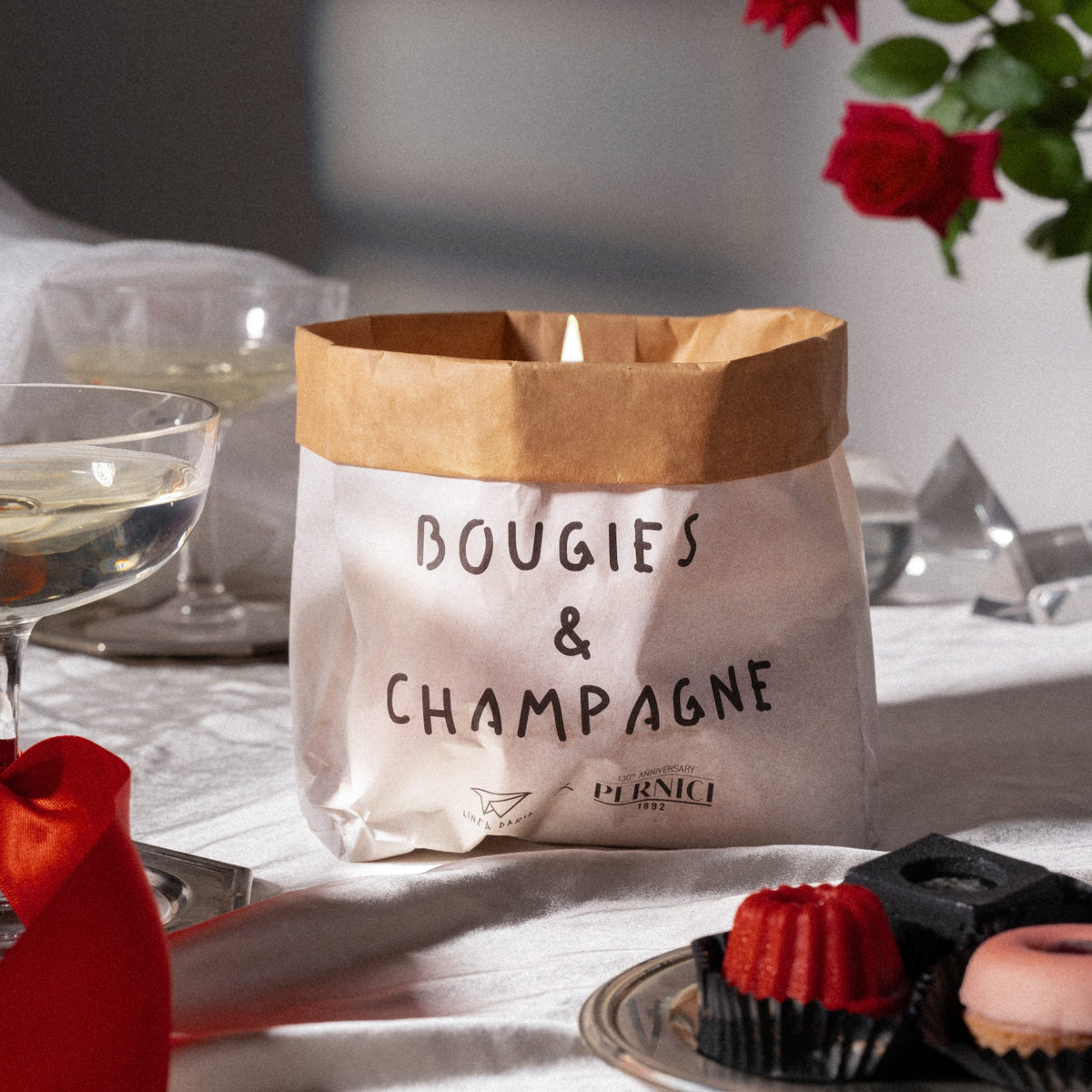 Candela profumata in sacchetto di carta con la scritta “Bougies & Champagne”, fragranza Talc & White Flower, ambientata su tavola raffinata con calici e dolci.